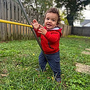 Albin is registered to the contest to win money with this photo: backyard, blue_pants, boots, child, curly_hair, daylight, fence, garden, grass, happy, outdoor, playful, playground, portrait, red_jacket, smiling, standing, swing, swing_set, toddler