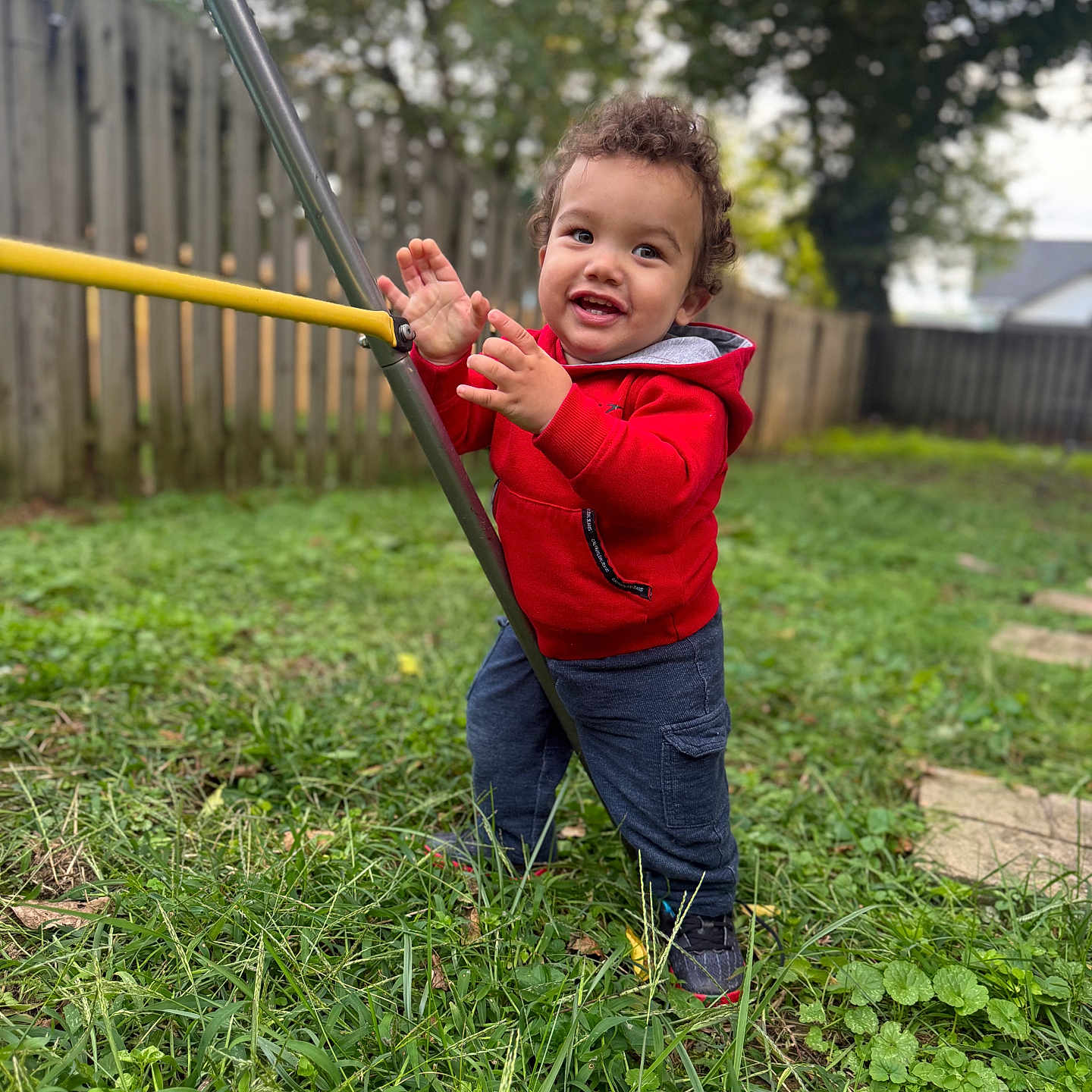 Albin is registered to the contest to win money with this photo: backyard, blue_pants, boots, child, curly_hair, daylight, fence, garden, grass, happy, outdoor, playful, playground, portrait, red_jacket, smiling, standing, swing, swing_set, toddler