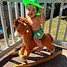 toddler, child, hat, green_hat, rocking_horse, toy, plush_toy, porch, sunlight, shadow, safety_net, wooden_railing, outdoor, summer, barefoot, shorts, baby, playtime, cute, smiling