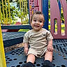 child, toddler, playground, smiling, curly_hair, outdoor, daylight, play_structure, yellow_railings, pink_slide, blue_platform, black_socks, short_sleeve_shirt, shorts, happy, sitting, nature, trees, grass, park