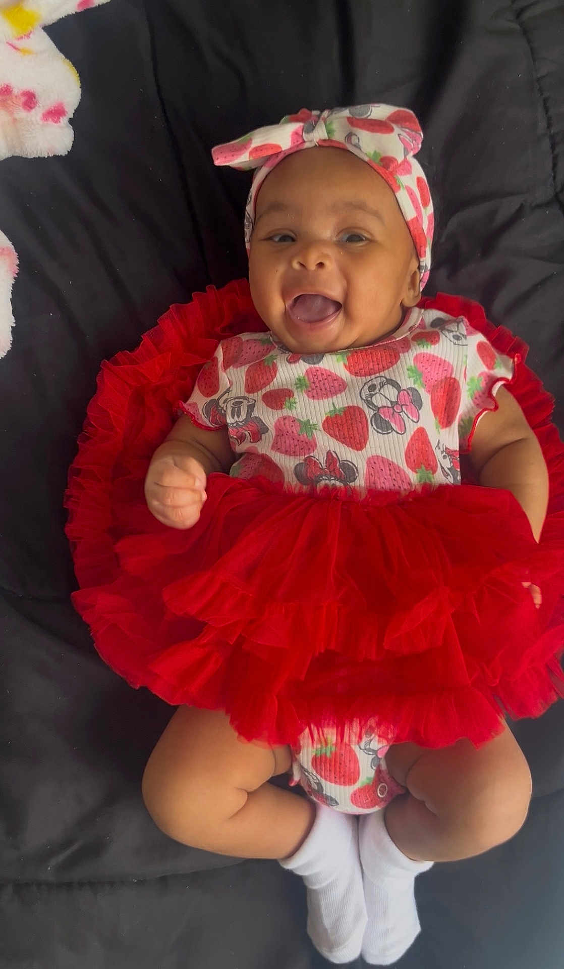 Kiara joined the competition — help win amazing prizes! baby, infant, smiling, tutu, headband, strawberry_pattern, red_clothing, white_socks, cute, happy, lying_down, child, adorable, tongue_out, black_background, playful, fashion, portrait, closeup, fun