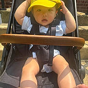 Haysen is registered to the contest to win money with this photo: baby, child, stroller, yellow_cap, sunlight, outdoor, brick_wall, steps, legs, feet, white_clothing, curious, hands, face, portrait, seatbelt, black_stroller, summer, cute, infant