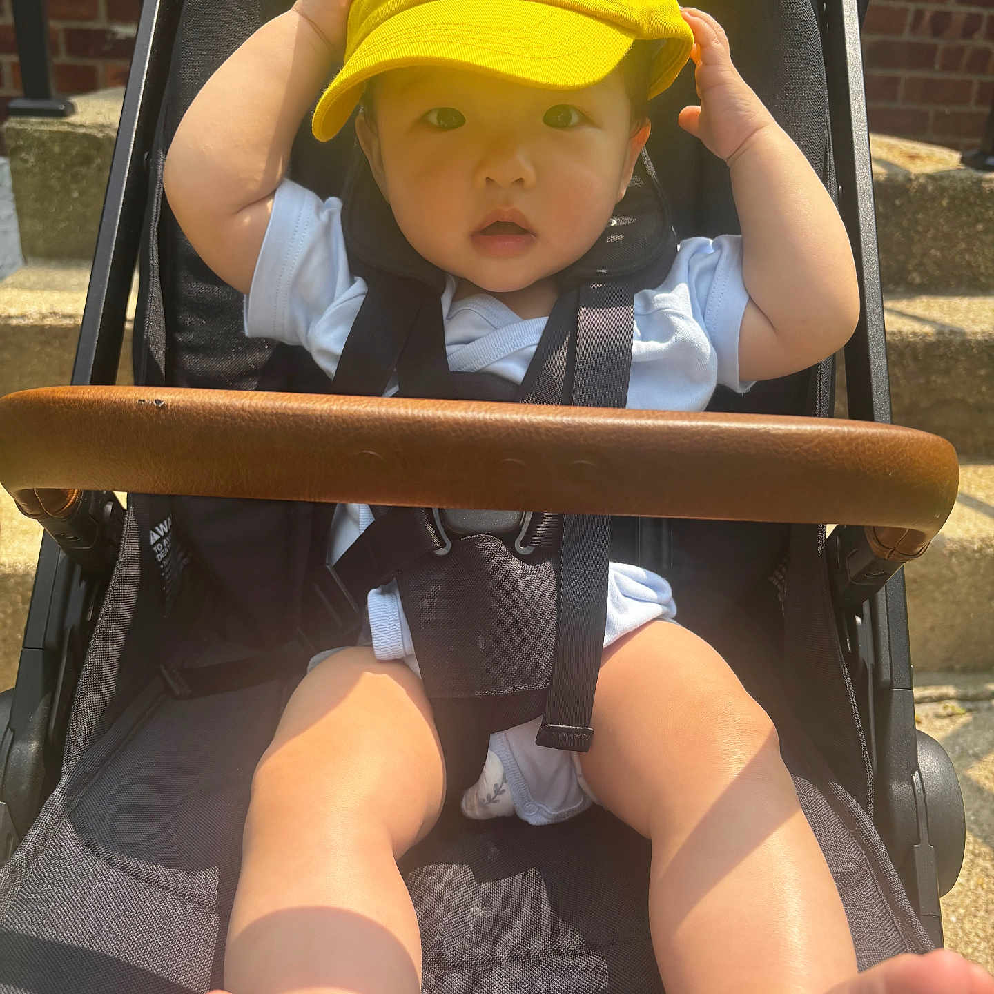 Haysen is registered to the contest to win money with this photo: baby, black_stroller, brick_wall, child, curious, cute, face, feet, hands, infant, legs, outdoor, portrait, seatbelt, steps, stroller, summer, sunlight, white_clothing, yellow_cap