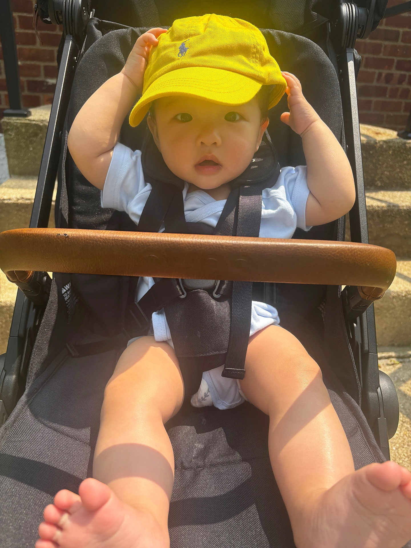 Haysen is registered to the contest to win money with this photo: baby, child, stroller, yellow_cap, sunlight, outdoor, brick_wall, steps, legs, feet, white_clothing, curious, hands, face, portrait, seatbelt, black_stroller, summer, cute, infant
