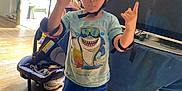 Daniel is registered to the contest to win money with this photo: child, helmet, knee_pads, tshirt, indoor, wooden_floor, television, toy_car_seat, colorful_socks, hand_gesture, blue_shorts, living_room, playful, safety_gear, person, casual_clothing, red_helmet, smiling, home_interior, furniture