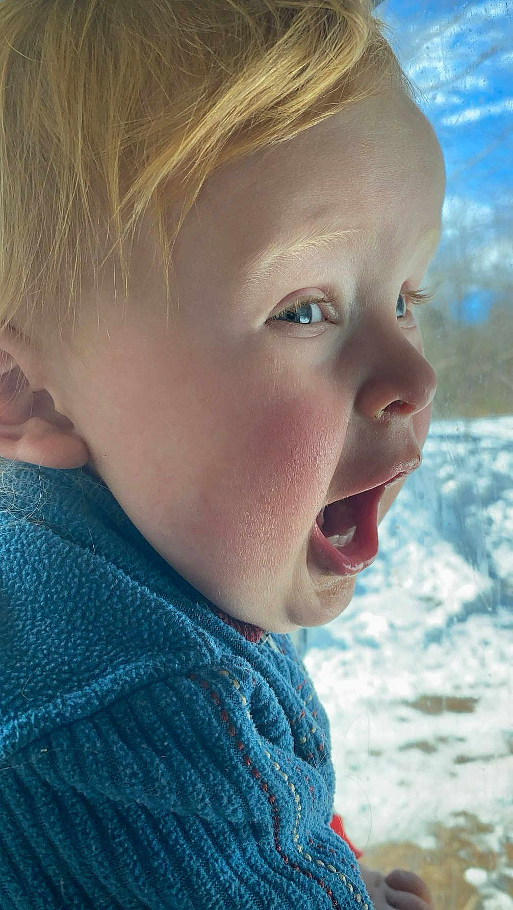 Abel Blake is registered to the contest to win money with this photo: child, blonde_hair, blue_eyes, side_profile, open_mouth, joyful_expression, blue_sweater, knitwear, window, snow, outdoor, winter, face, skin, eyelashes, ear, nose, cheeks, young, portrait