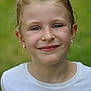 child, girl, smile, face, blue_eyes, blonde_hair, earrings, white_shirt, paint_stains, outdoor, green_background, portrait, young, casual_clothing, natural_light, close_up, happy, primary_school, headshot, expression