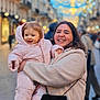 child, toddler, woman, smiling, happy, winter_clothing, pink_snowsuit, street, blurred_background, festive_lights, outdoor, urban, holding, joy, cozy, cold_weather, people, walking, bag, portrait