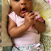 Jamoni is registered to the contest to win money with this photo: baby, infant, headband, bow, cute, expression, hands, clasped, pink_shirt, floral_shorts, quilt, blanket, indoor, portrait, child, person, sleepwear, smiling, closeup, cozy