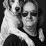 adult, black_and_white, black_shirt, close_up, companionship, dark_background, dog, dramatic_lighting, floppy_ears, golden_retriever, long_hair, owner, paws, person, pet, portrait, shoulder, studio, sunglasses, white_fur