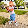 toddler, child, smiling, outdoors, sidewalk, grass, tree, neighborhood, houses, cars, parked_cars, toy, red_toy, white_shirt, blue_shorts, sneakers, nike, standing, portrait, daylight