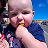 Kyler is registered to the contest to win money with this photo: baby, child, lollipop, outdoor, blue_sky, sunlight, face, hand, clothing, short_sleeve, cute, person, summer, daytime, street, building, sunglasses, graphic_tee, skin, closeup