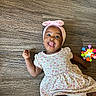 baby, child, floor, wooden_floor, headband, pink, bow, dress, floral_pattern, toy, colorful, tongue_out, happy, smiling, infant, person, cute, indoors, playful, sitting
