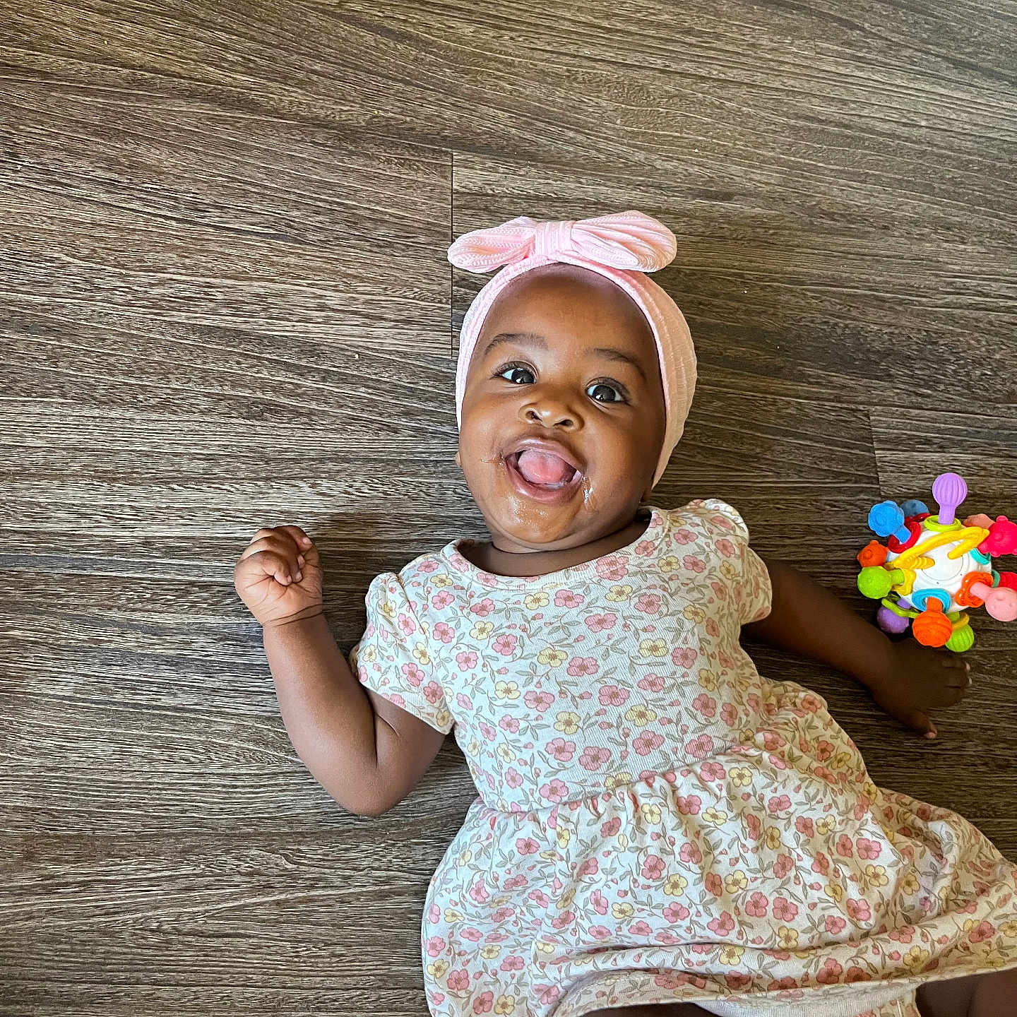 Malaysia Mells is registered to the contest to win money with this photo: baby, child, floor, wooden_floor, headband, pink, bow, dress, floral_pattern, toy, colorful, tongue_out, happy, smiling, infant, person, cute, indoors, playful, sitting