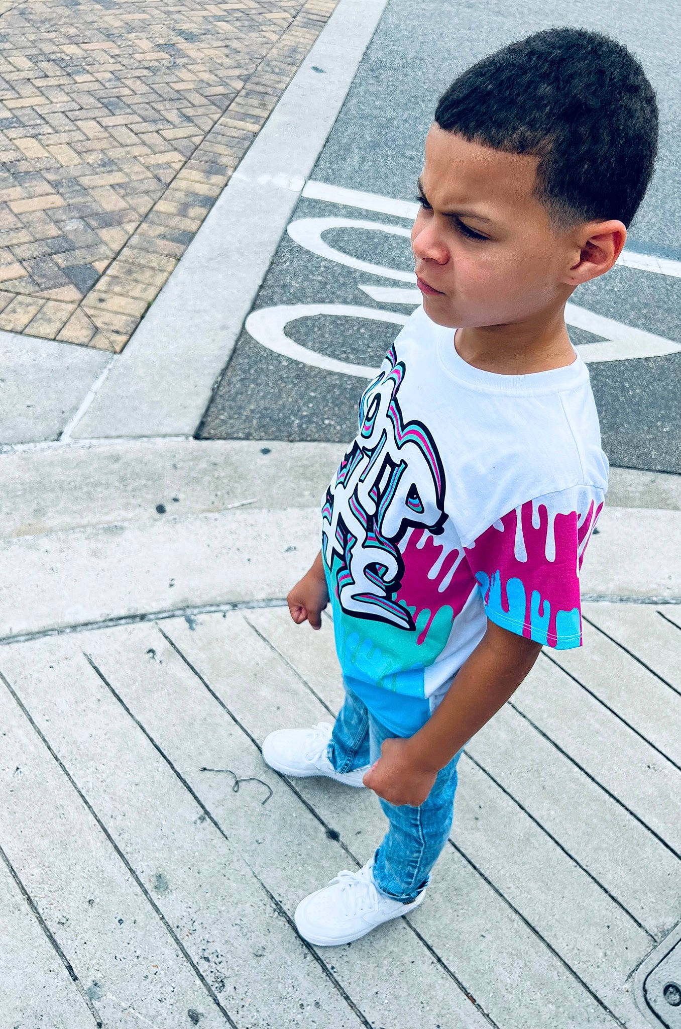 Kingston is registered to the contest to win money with this photo: asphalt, baby_toddler_clothing, black_hair, child, cool, electric_blue, face, flooring, happy, head, human_leg, leisure, pattern, person, sandal, sitting, sleeve, t_shirt, thigh, toddler