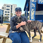 Mulan is registered to the contest to win money with this photo: person, dog, bench, outdoor, cap, smile, building, sky, wooden_bench, casual_clothing, pet, canine, daylight, happy, animal, urban, friendship, leash, brindle, human