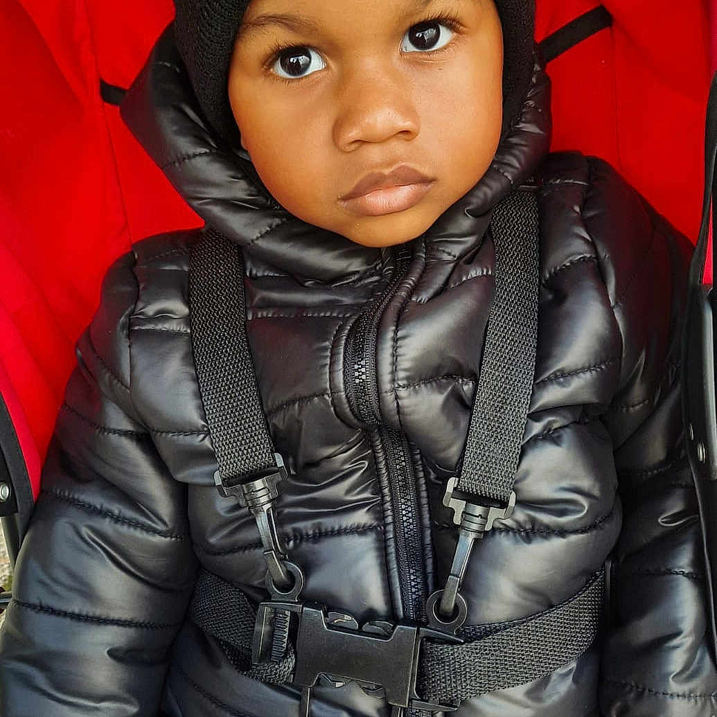 Gabriel participe au concours pour gagner de l'argent avec cette photo : baby, beanie, black_jacket, blue_pants, child, clothing, dinosaur_logo, face, hand, hat, outerwear, patterned_pants, portrait, puffer_jacket, red_stroller, seatbelt, serious_expression, straps, stroller, toddler