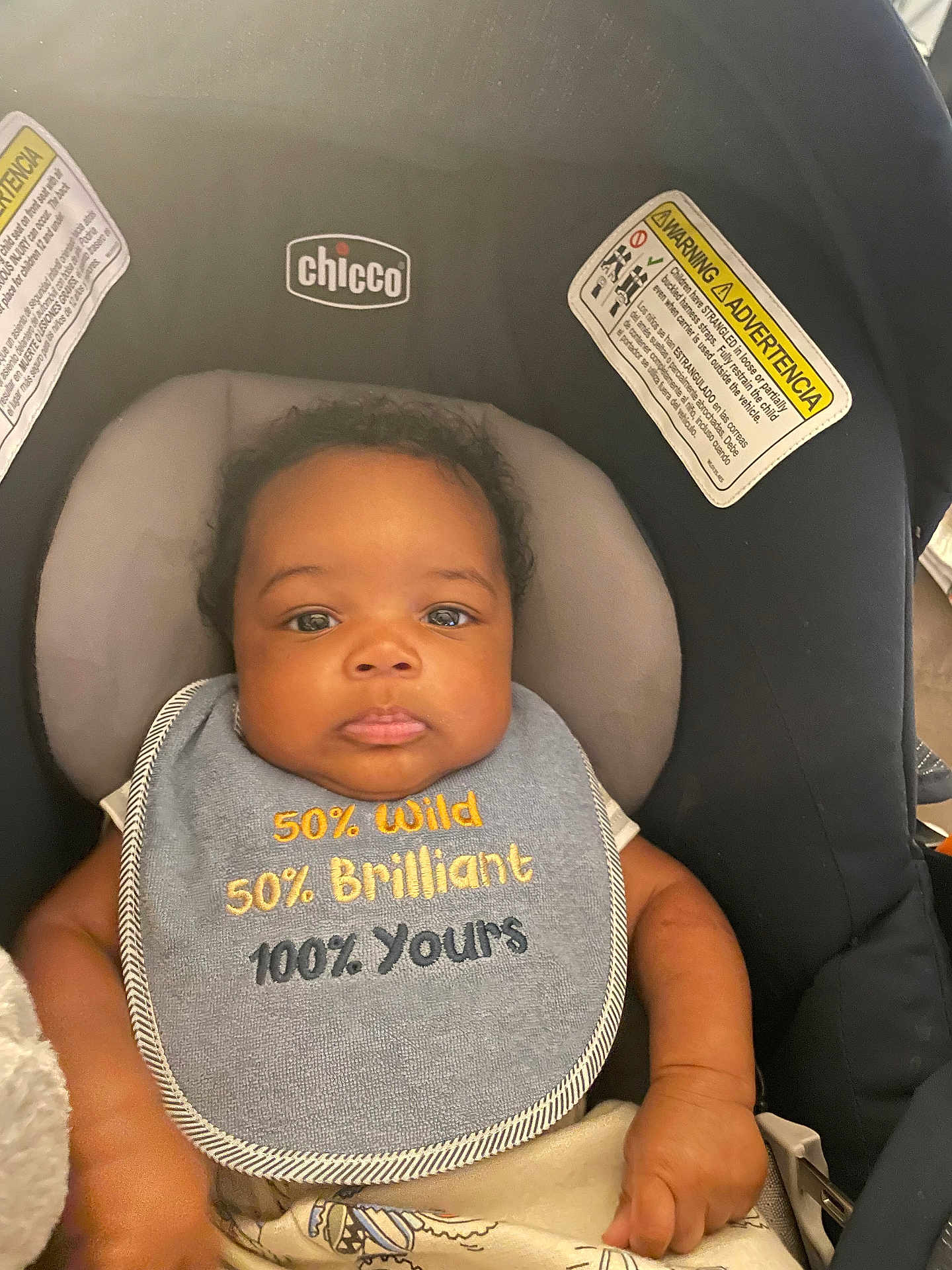 Lenox joined the competition — help win amazing prizes! baby, infant, car_seat, chicco, bib, clothing, face, child, cute, serious_expression, text_on_clothing, seatbelt, safety_warning, indoor, person, head, arm, skin, young_child, closeup