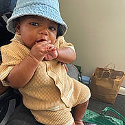 Lenox is registered to the contest to win money with this photo: baby, child, denim_hat, bucket_hat, knit_outfit, beige_clothing, hand_in_mouth, sitting, indoor, person, curious_expression, brown_skin, shorts, buttoned_shirt, foot, bag, paper_bag, green_bag, casual, relaxed