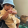 baby, child, denim_hat, bucket_hat, knit_outfit, beige_clothing, hand_in_mouth, sitting, indoor, person, curious_expression, brown_skin, shorts, buttoned_shirt, foot, bag, paper_bag, green_bag, casual, relaxed