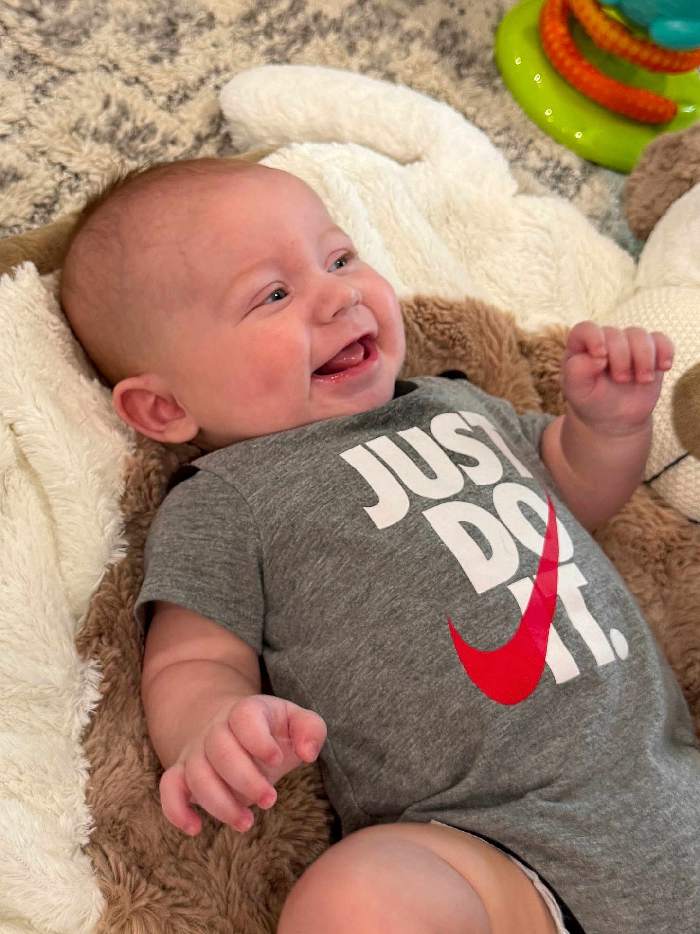 Isaiah joined the competition — help win amazing prizes! baby, infant, onesie, smiling, laughing, plush_blanket, toy, carpet, hand, face, cheeks, lying_down, gray_clothing, slogan_text, red_swoosh, happy, indoor, soft_texture, portrait, cute
