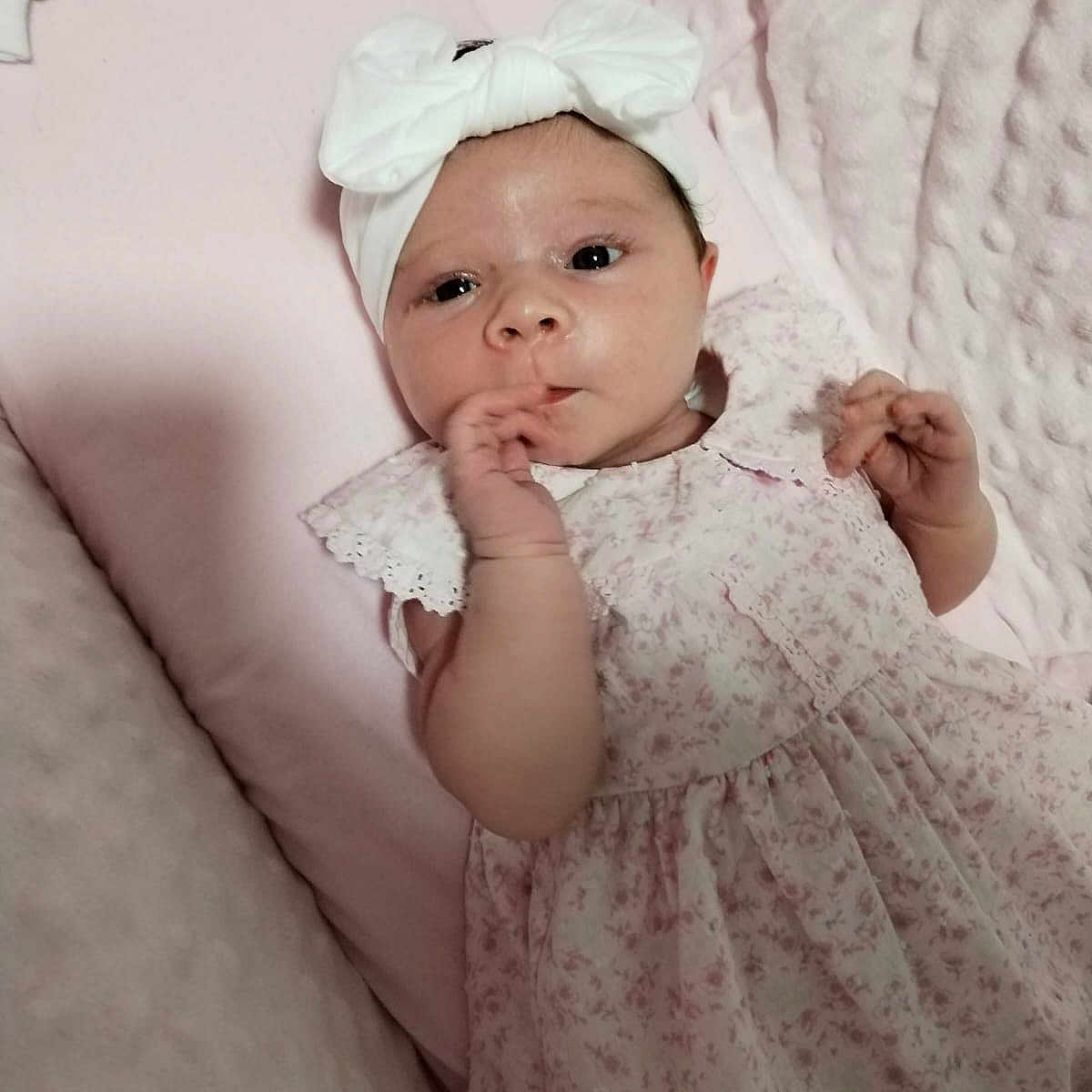 Isla joined the competition — help win amazing prizes! baby, blanket, bow, child, cute, expression, face, floral_dress, girl, hand_near_mouth, headband, indoors, infant, lying_down, newborn, pink, portrait, skin, soft_texture, toddler