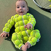 Emily is registered to the contest to win money with this photo: baby, tennis_balls, tennis_court, tennis_racket, smiling, infant, outdoor, playful, sportswear, unique_costume, child, green, recreation, cute, feet, hands, face, young, fun, leisure