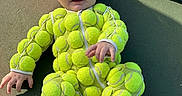 Emily is registered to the contest to win money with this photo: baby, child, tennis_ball, tennis_racket, tennis_court, green, outdoor, sportswear, cute, infant, footwear, smiling, playful, one_person, sitting, clothing, face, hands, feet, shadow