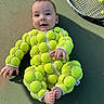baby, child, tennis_ball, tennis_racket, tennis_court, green, outdoor, sportswear, cute, infant, footwear, smiling, playful, one_person, sitting, clothing, face, hands, feet, shadow