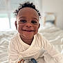 baby, smiling, curly_hair, white_clothing, toy, teddy_bear, bed, blanket, indoors, child, happy, cute, portrait, person, infant, face, closeup, soft_light, comfort, playtime