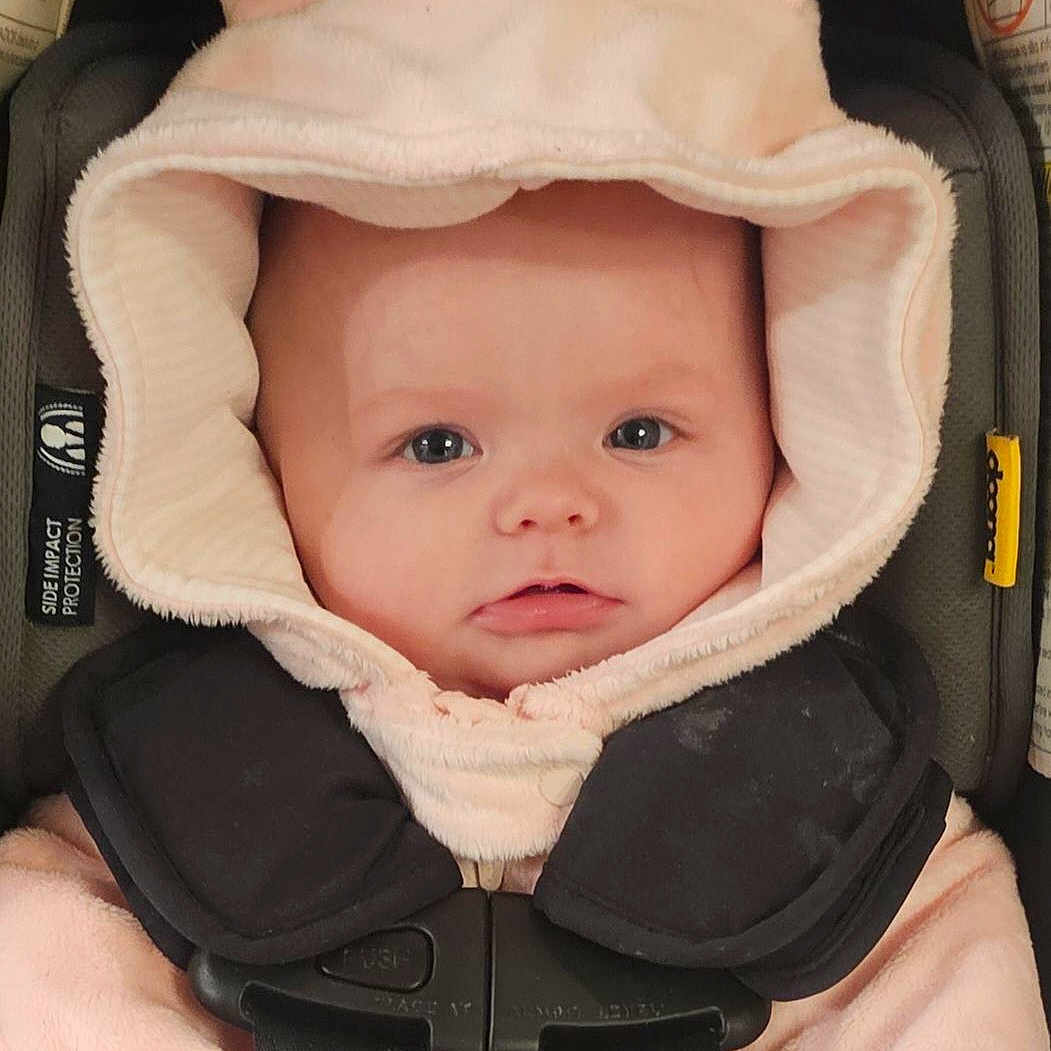 Scotland is registered to the contest to win money with this photo: apparel, baby, bunny_hoodie, car_seat, child, closeup, cute, expression, face, hood, indoors, infant, person, pink_clothing, portrait, safety, seatbelt, soft_texture, straps, young_child