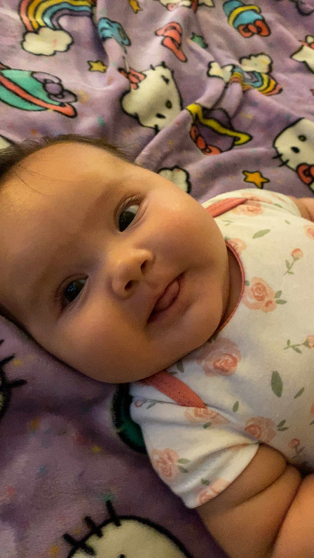 Ariya is registered to the contest to win money with this photo: baby, infant, face, tongue, blanket, hello_kitty, pattern, cute, child, lying_down, purple, smiling, clothing, floral, onesie, soft, playful, closeup, indoors, person
