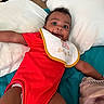 Zoya is registered to the contest to win money with this photo: baby, infant, red_onesie, bib, smile, pillow, bed, blanket, happy, child, indoors, person, cute, lying_down, face, skin, head, expression, young, cozy