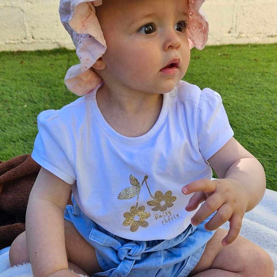 Ambre a rejoint le concours — aidez-le/la à gagner de superbes lots ! baby, blanket, casual_clothing, child, curious, cute, daylight, golden_flowers, grass, hat, infant, nature, outdoor, pink_hat, portrait, sitting, soft_texture, sunhat, white_shirt, young_child