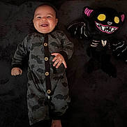 Elias is registered to the contest to win money with this photo: baby, bat_pattern, black_background, child, costume, cute, dark_colors, happy, indoor, infant, infant_clothing, lying_down, onesie, playful, plush, portrait, smiling, soft_texture, stuffed_toy, toy