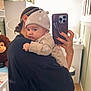 baby, adult, mirror, selfie, phone, hat, plush_toy, nursery, room, person, clothing, hand, furniture, wall, decor, child, holding, portrait, indoor, soft_light