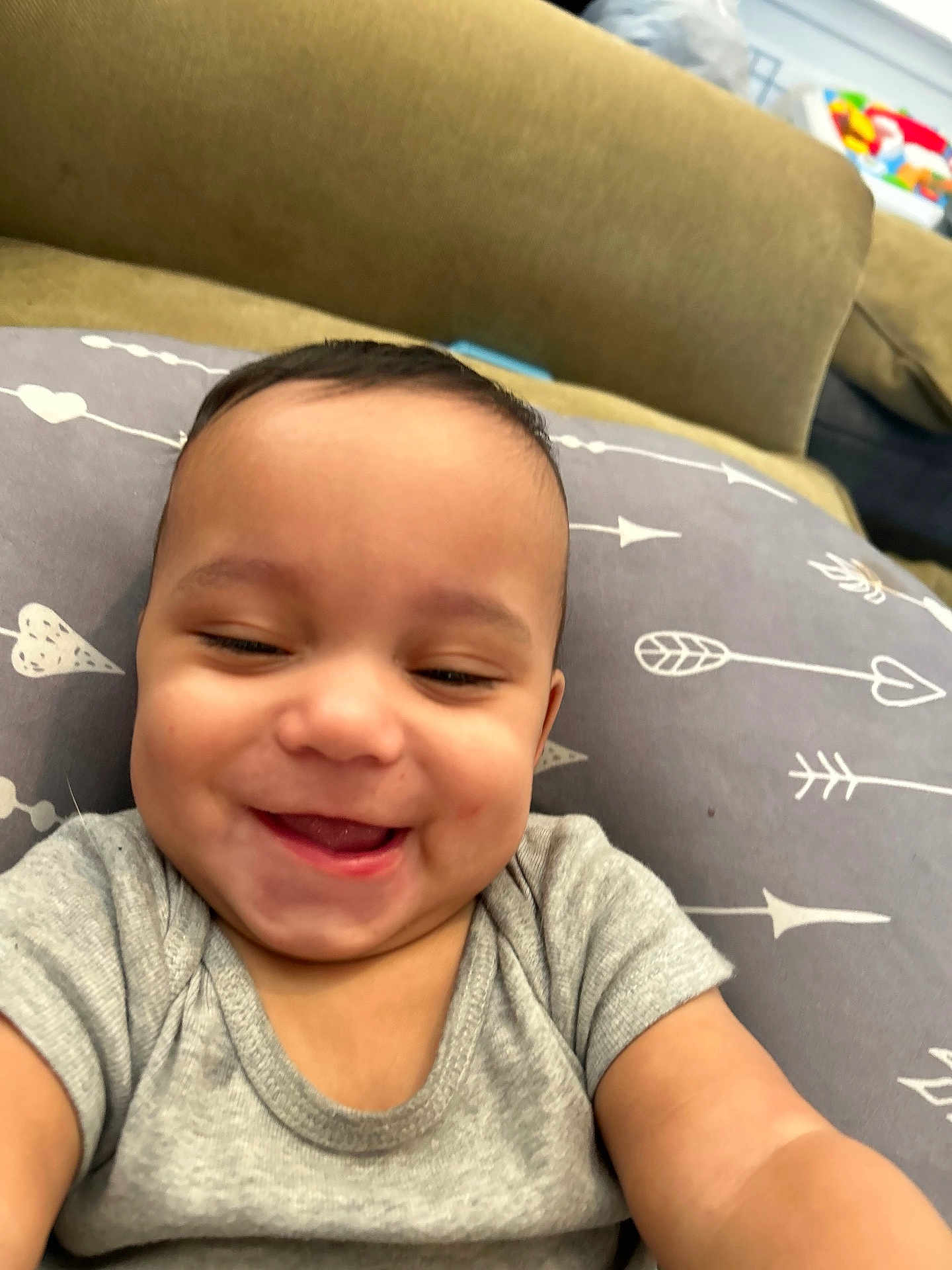 Kaiden is registered to the contest to win money with this photo: baby, smiling, infant, pillow, arrows, hearts, clothing, gray_shirt, cute, happy, face, child, indoors, closeup, person, skin, head, expression, joy, young