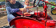 Kaiden joined the competition — help win amazing prizes! baby, child, woman, tractor, red, outdoor, farm, vintage, steering_wheel, vehicle, person, sunny, daytime, blue_sky, tree, clothing, casual, smile, playing, agriculture
