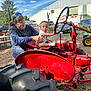 baby, child, woman, tractor, red, outdoor, farm, vintage, steering_wheel, vehicle, person, sunny, daytime, blue_sky, tree, clothing, casual, smile, playing, agriculture