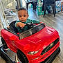 Kaiden is registered to the contest to win money with this photo: toddler, child, toy_car, indoor, flooring, steering_wheel, red, black, chair, table, box, water_pack, window, daylight, person, floor, plastic, home, baby_toy, furniture