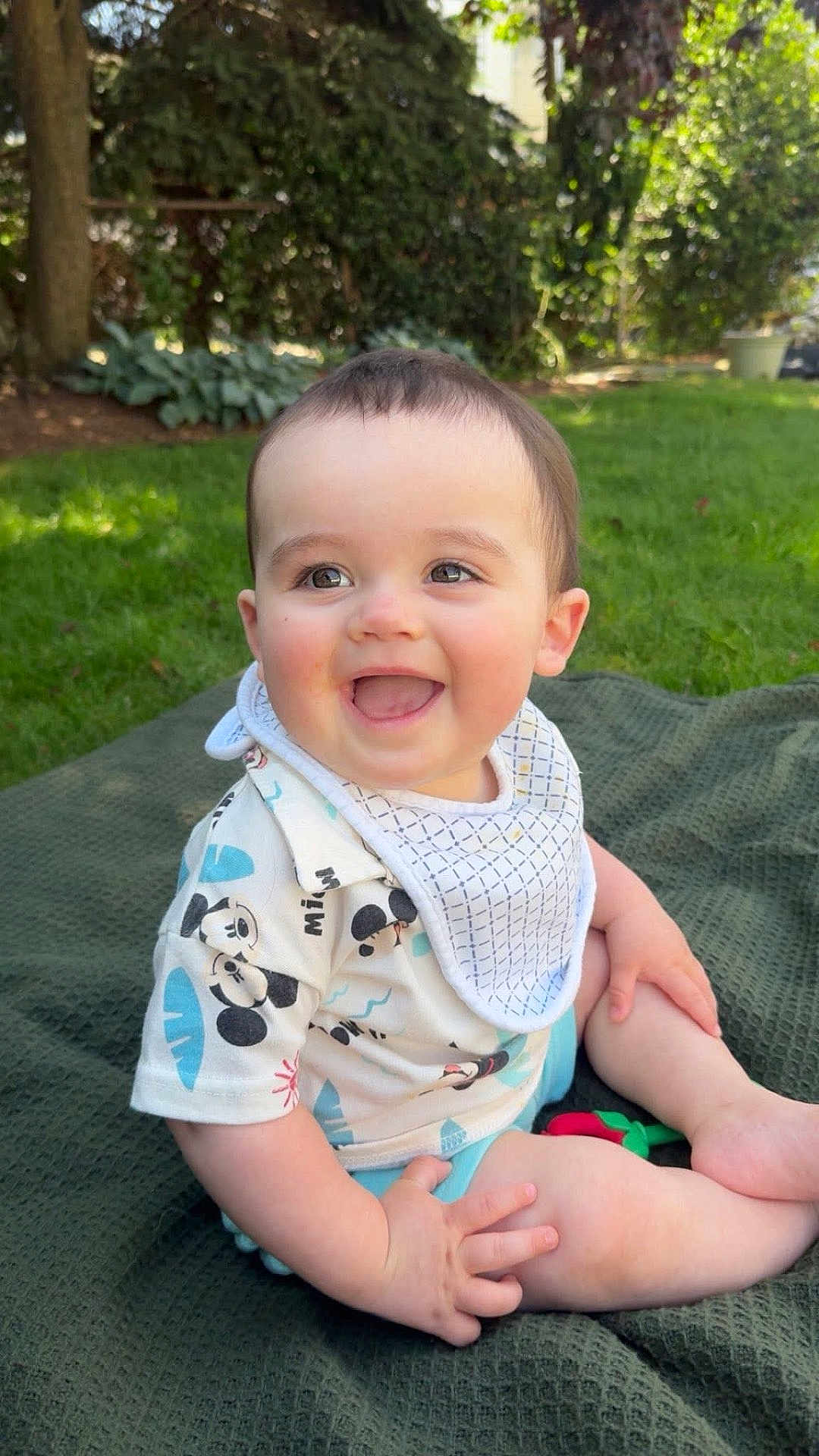Declan joined the competition — help win amazing prizes! baby, child, smiling, outdoor, grass, blanket, bib, shirt, mickey_mouse, happy, cute, sitting, nature, greenery, sunlight, infant, portrait, face, cheerful, daylight