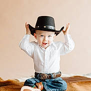 Jasper is registered to the contest to win money with this photo: beige_background, belt, belt_buckle, blue_eyes, child, cowboy_boots, cowboy_hat, cute, fur_blanket, hands_up, hat, indoor, jeans, person, playful, portrait, sitting, smile, toddler, white_shirt