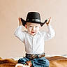 beige_background, belt, belt_buckle, blue_eyes, child, cowboy_boots, cowboy_hat, cute, fur_blanket, hands_up, hat, indoor, jeans, person, playful, portrait, sitting, smile, toddler, white_shirt