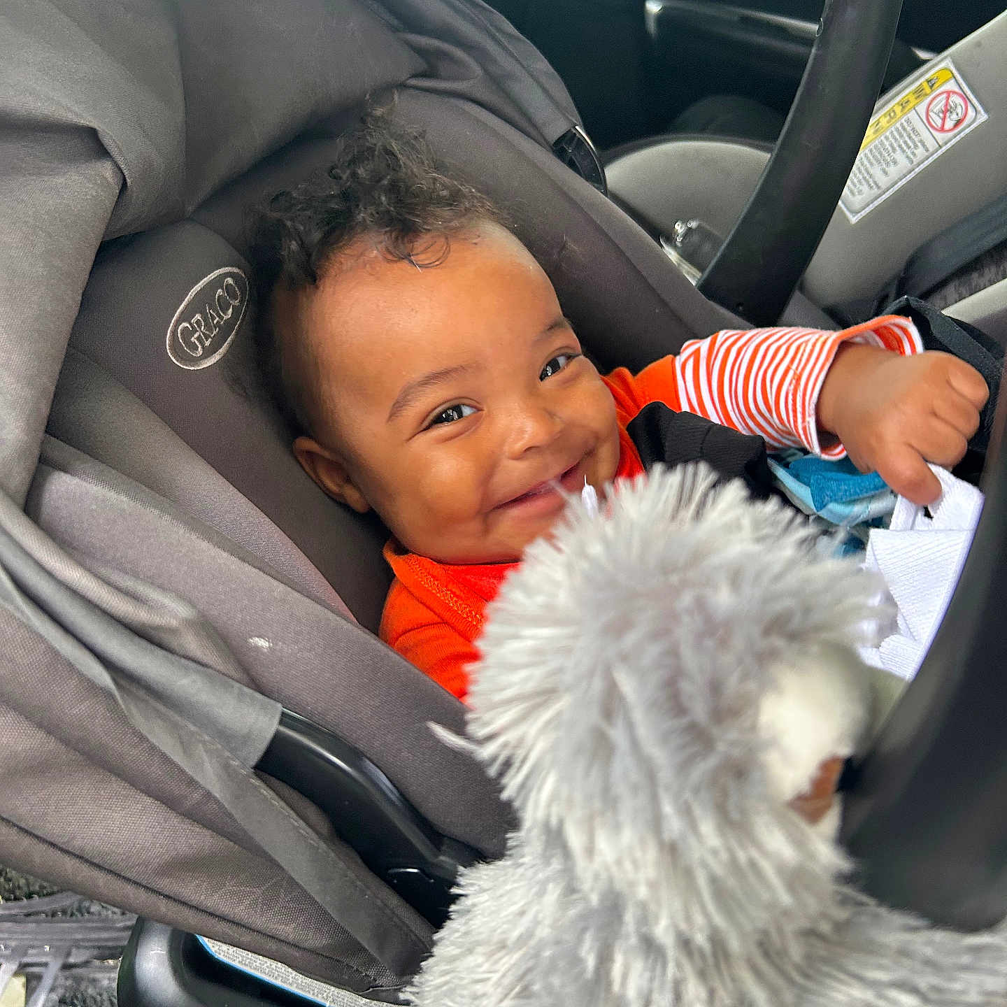 Zhyaire is registered to the contest to win money with this photo: baby, car_interior, car_seat, child, curly_hair, cute, gray, happy, indoors, infant, orange_clothing, person, playful, plush, portrait, seatbelt, smile, stuffed_animal, toy, young_child