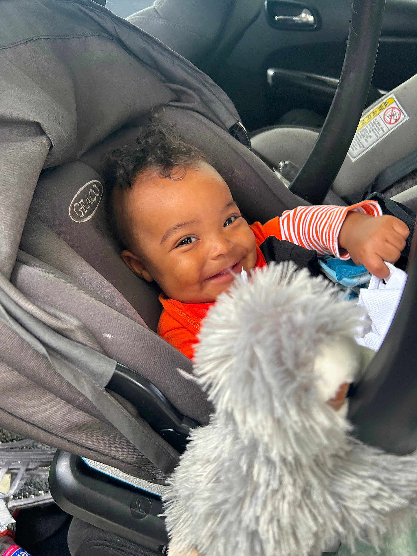 Zhyaire is registered to the contest to win money with this photo: baby, car_seat, smile, child, toy, stuffed_animal, gray, orange_clothing, infant, seatbelt, happy, car_interior, person, cute, plush, portrait, young_child, curly_hair, indoors, playful