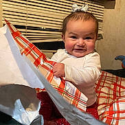 Ariella is registered to the contest to win money with this photo: baby, child, smiling, happy, indoor, wrapping_paper, gift, red_pants, white_shirt, bow, window_blind, curtain, face, hands, sitting, cheeks, excitement, cozy, person, portrait