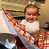 baby, child, smiling, happy, indoor, wrapping_paper, gift, red_pants, white_shirt, bow, window_blind, curtain, face, hands, sitting, cheeks, excitement, cozy, person, portrait