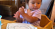 Ariella joined the competition — help win amazing prizes! toddler, child, high_chair, crayon, drawing, bird, table, restaurant, indoor, wooden_chair, clothing, person, coloring, artwork, hair_tie, face, expression, booth_seating, lighting, casual