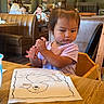 toddler, child, high_chair, crayon, drawing, bird, table, restaurant, indoor, wooden_chair, clothing, person, coloring, artwork, hair_tie, face, expression, booth_seating, lighting, casual