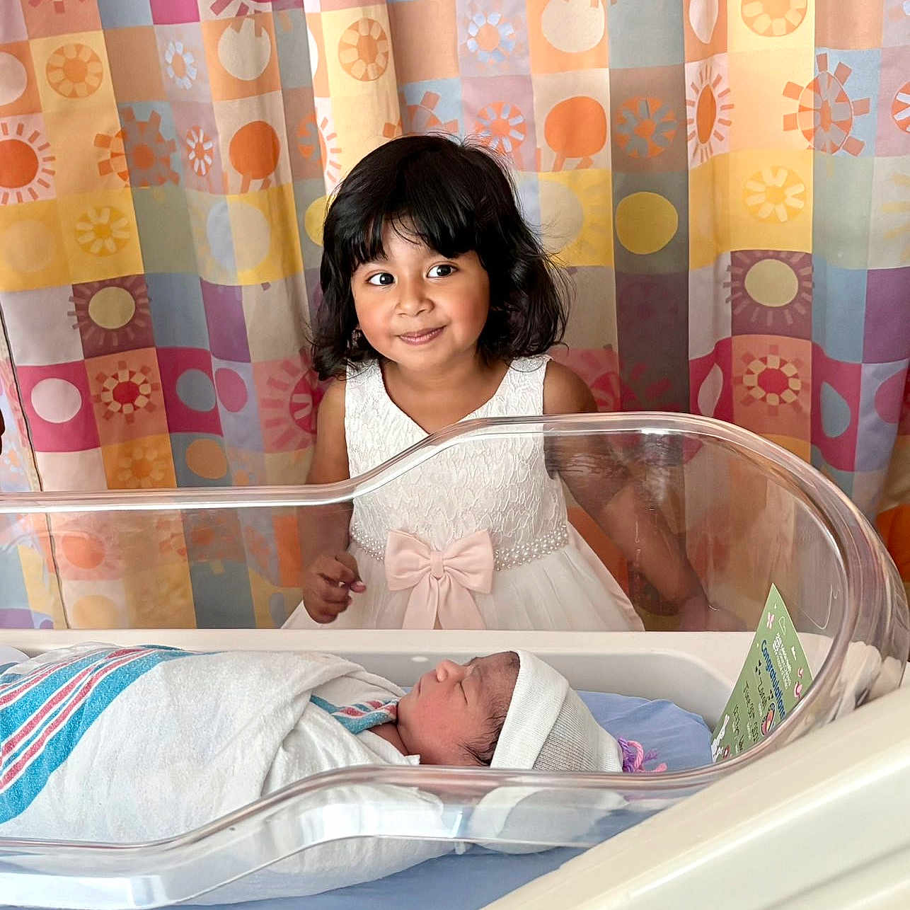 Sarah is registered to the contest to win money with this photo: baby, newborn, child, girl, hospital, bassinet, blanket, curtain, pattern, dress, bow, smile, portrait, indoor, person, infant, hospital_room, cute, happy, family